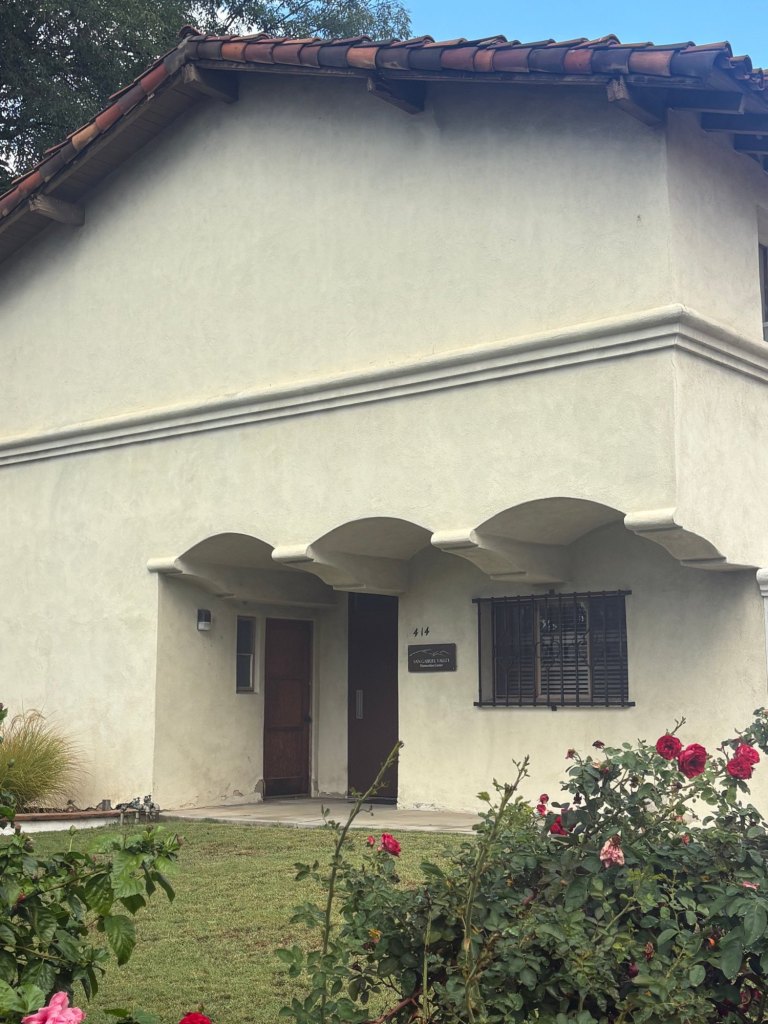 The front of the Center's building at 414 South Mission Drive: a white stucco Mission Revival building with a red tile roof, arched overhangs above the entry, and the Center's sign beside the door, with rose bushes in the foreground.
