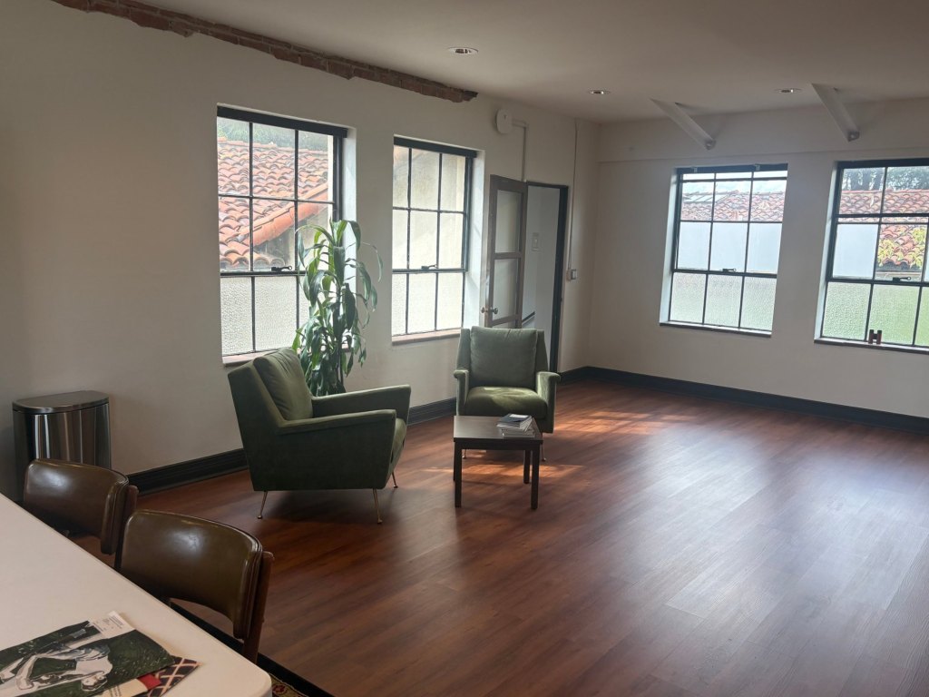 A sunlit room at the Center with two green armchairs, a small table, and a tall plant beside steel-framed windows overlooking red tile roofs.